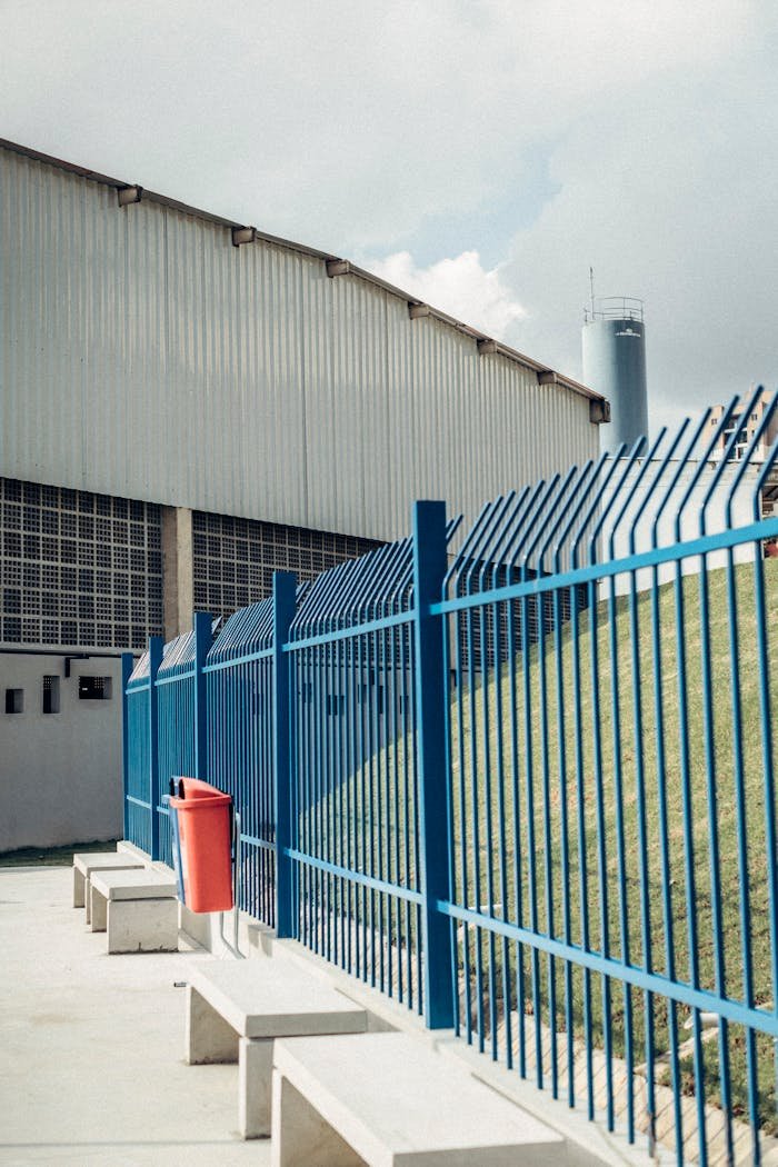 An industrial warehouse features a blue security fence and stone benches under a clear sunny sky.